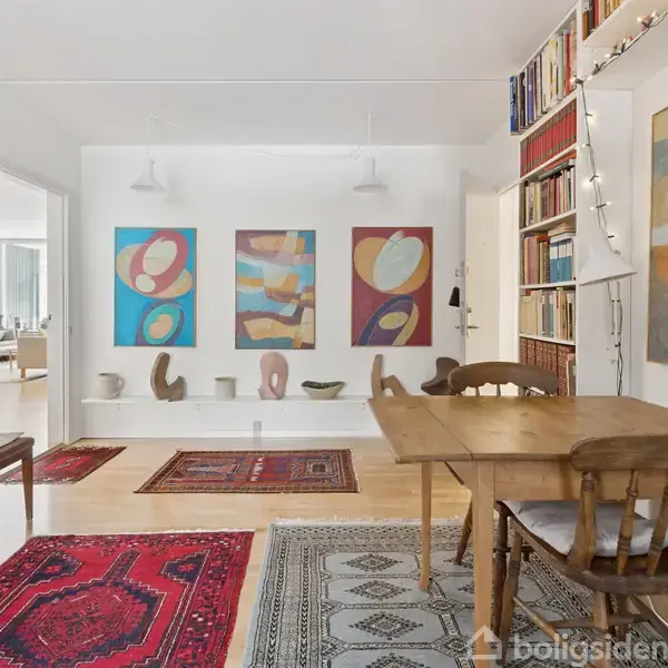 Træbord og stol står i en stue med tæpper og kunst på væggene. Hylder fyldt med bøger til højre, abstrakte malerier hænger foran, suppleret af forskellige kunstgenstande på gulvet.