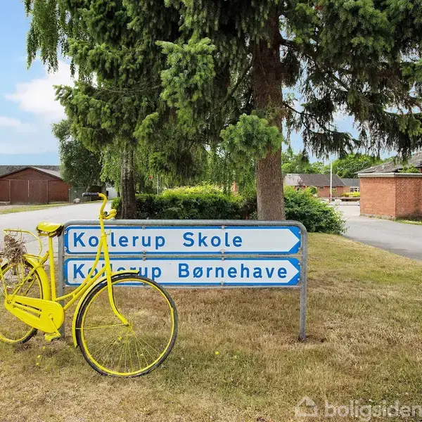 Gul cykel læner sig op ad vejskilte vedrørende "Koltrup Skole" og "Koltrup Børnehave" på en græsplæne under et grantræ. Baggrunden viser huse og en vej i et boligområde.
