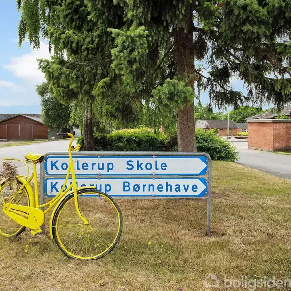 En gul cykel står op ad et vejskilt med teksten "Kollerup Skole" og "Kollerup Børnehave." Skiltet er placeret ved siden af en vej under et grantræ i et boligområde.
