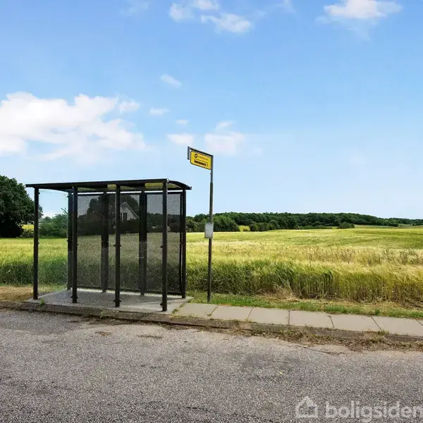 En glasbusstoppested står ved en landevej, omgivet af grønne marker og en blå himmel med få skyer. Et busstoppestedsmærke er placeret ved siden af det.