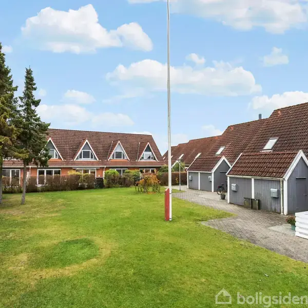 En række af murstenshuse med røde tage står i en gård. Foran husene er der en græsplæne og en flagstang. To høje træer ses til venstre.