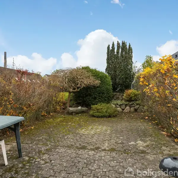 En stenbelagt terrasse med et bord og en stol står omgivet af buske og træer. Terrasse fører til en have med klippede buske og en hæk. Himlen er blå med skyer.