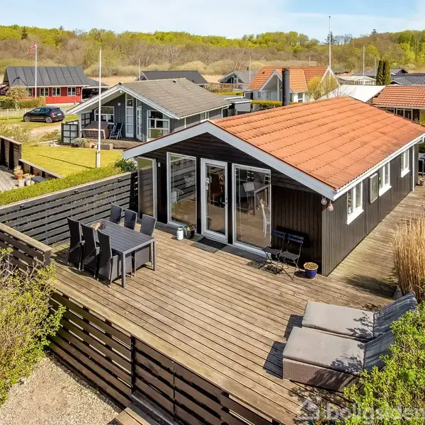 Sort træsommerhus med rødt tag har en stor træterrasse med havemøbler. Det er placeret i et sommerhusområde med flere huse og grønne træer i baggrunden.
