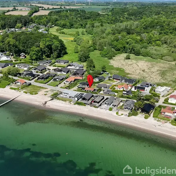 En strand ved en række villaer med skov og marker i baggrunden. En rød markør er placeret over en af villaerne, der ligger tæt ved kysten.