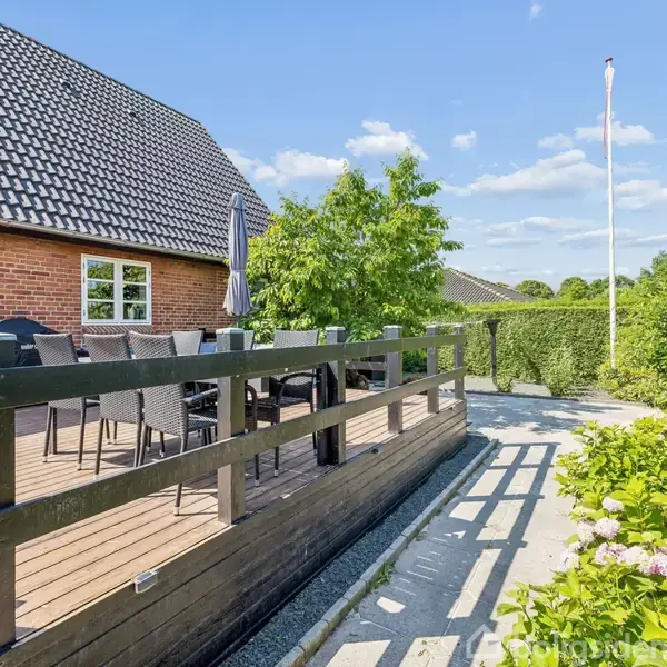 Terrasse med sorte havemøbler ved siden af et rødstenshus, omgivet af grønne buske og træer. Blå himmel og enkelte skyer i baggrunden. Grussti med små blomster langs kanten.
