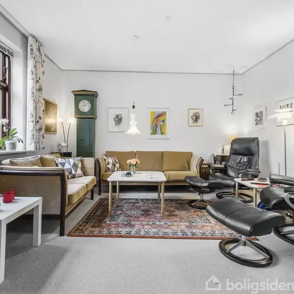 Stue med sofa og lænestole omkring sofabord, tæppe under. Væg med malerier, ur i hjørnet. Vindue med planter, lysindfald. Belysning fra lamper. Hyggelig atmosfære.