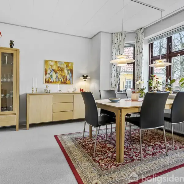 Et spisebord med fire stole står på et orientalsk tæppe i en lys spisestue. Store vinduer med gardiner omkranser rummet. En skænk og vitrineskab findes i hjørnet.