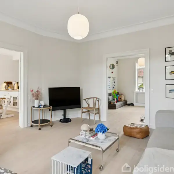 Stue med sofa og fjernsyn, lysmalet væg med døre til tilstødende rum. Der er en blanding af moderne og stilfuldt interiør samt lysindfald fra vinduer i baggrunden.