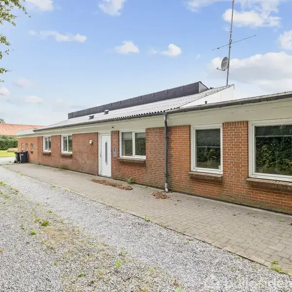 En lang, enetages murstensbygning med hvide vinduer og en hvid dør. Bygningen ligger ved en grusindkørsel med grønne træer og en åben himmel i baggrunden. Skilt ved huset: "Fysioterapeut Birgit Præst".