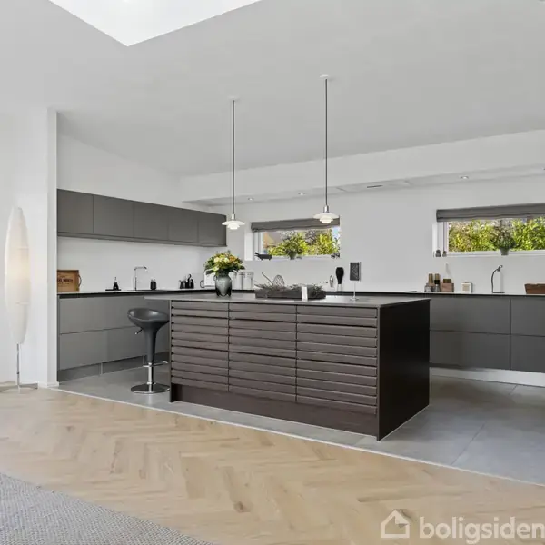 Moderne køkken med en stor, mørk køkkenø omgivet af barstole. Indbygningspejs til højre brænder lyst. Lyse trægulve, store vinduer med udsigt til have. Ingen tekst.