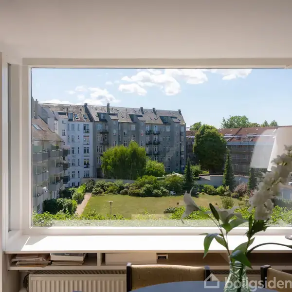 Stort vindue åbner sig mod en udsigt over en grøn gård omkranset af lejlighedsbygninger. En hvid lampe og en vase med blomster står på en rund bordplade i forgrunden.