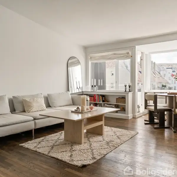 Sofaen står ved siden af et lavt sofabord på et tæppe, mens spisebordet er i baggrunden nær store vinduer, der giver udsigt til bygninger udenfor.
