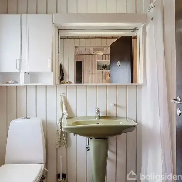Toilet og vask står foran en spejl og cremefarvede vægge i et badeværelse, omgivet af et kabinet og en dør.