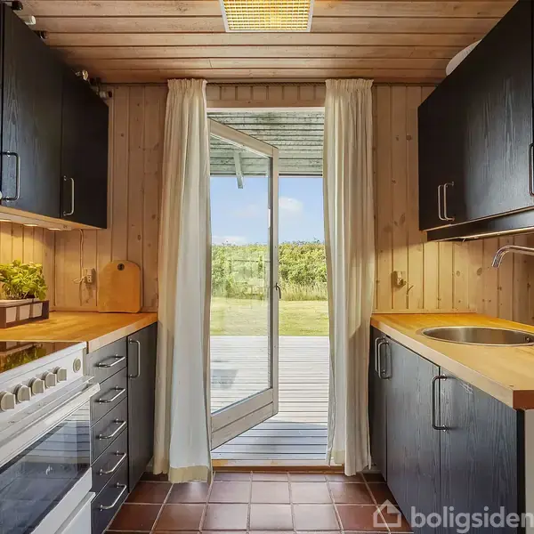 Et køkken med sorte skabe og træbordplader åbner ud til en terrasse gennem en glasdør. Rummet har trævægge og et flisegulv. Curtains hænger ved døren.