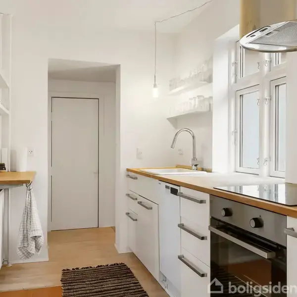 Et moderne køkken med hvide skabe og træbordplader. En ovn og kogeplade er integreret i enheden. Et vindue giver naturligt lys, mens en tæppe løber ligger på gulvet.