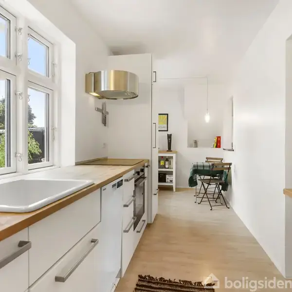 Køkken med hvidt interiør, træbordplader og vindue over vask. Indbyggede skabe og moderne emhætte. I baggrunden ses en spiseplads med et lille bord og stole.