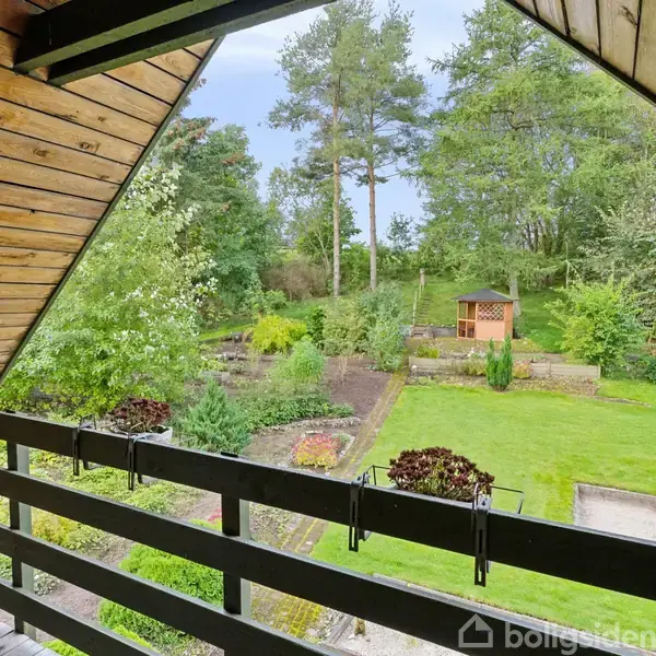 En terrasse med sort rækværk strækker sig ud over en velplejet have med grønne plæner, blomsterbede og træer. I baggrunden ses en lille træpavillon omgivet af natur.