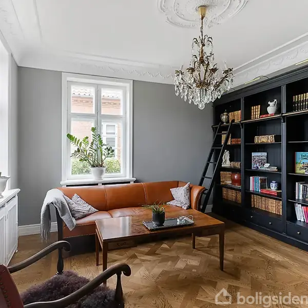 En stue med en orange sofa foran to vinduer. En stor bogreol fyldt med bøger og dekorationer står til højre med en stige ved siden af. Trægulv og elegant lysekrone.