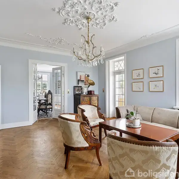 Stue med antikke møbler og lysekrone, arrangeret omkring et træbord. Lyse vægge og loftsudsmykning skaber en elegant atmosfære med vinduer, der giver udsigt til haven.