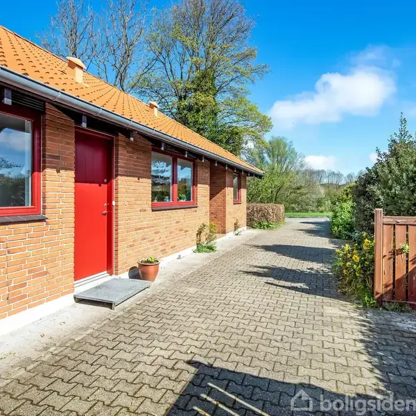 Et murstenshus med rødt tag og røde vinduer står i sollys. En flisebelagt sti fører forbi huset. Baggrunden er en have med træer og buske.