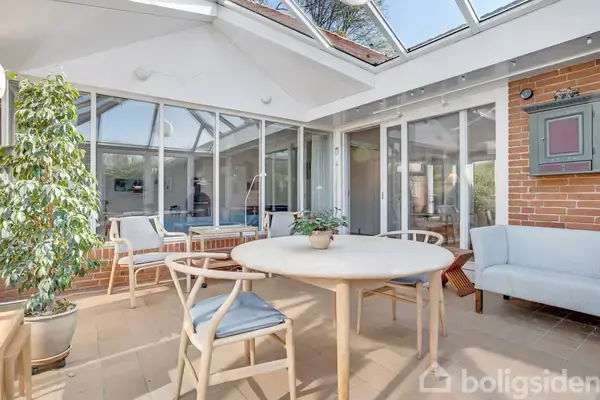 Et lyst rum med et rundt træbord omgivet af stole, planter ved siden af, og det hele i et glashus med udsigt til himlen, som skaber en naturlig atmosfære.