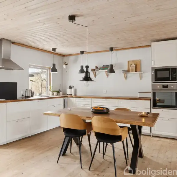 Køkkenbord står med fire stole omkring sig, pyntet med en skål. Køkkenet er lyst med træloft og moderne hvide skabe. En ovn og mikroovn er indbygget i skabene til højre.