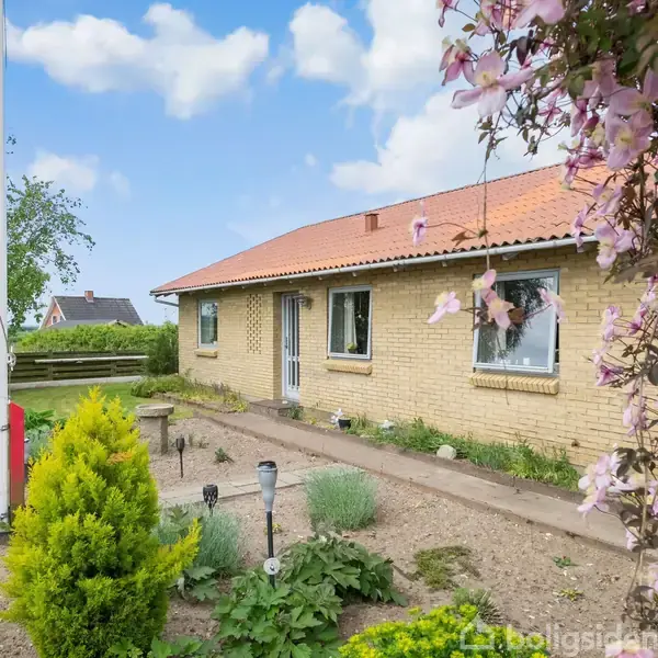 Et gult murstenshus med rødt tegltag står blandt blomstrende buske og velplejede bede i en have. Blå himmel med hvide skyer danner baggrund. Ingen tekst er til stede.