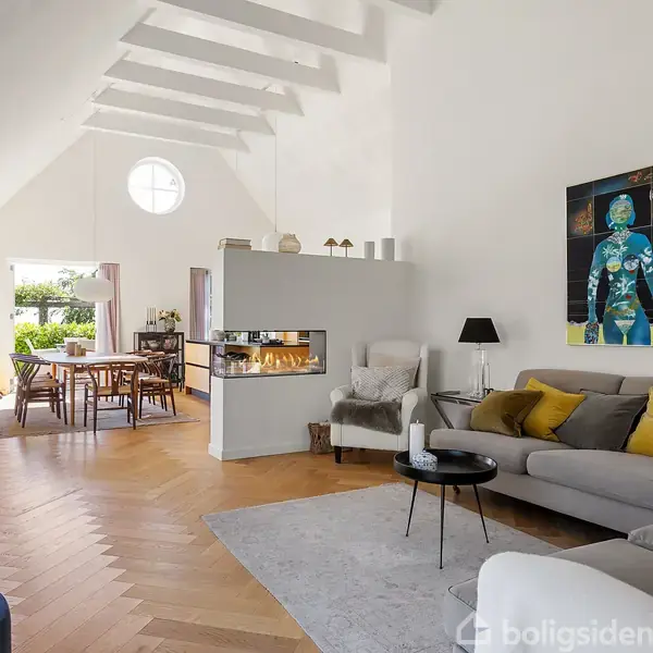 Stue med sofagruppe foran moderne pejs, lyse trægulve og kunst på væggen. Lyst spiseområde med store vinduer i baggrunden, som åbner ud mod en have.