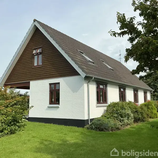 Et hvidt, trekantet hus står roligt omgivet af grønne buske og en velplejet græsplæne, med et mørkt skråt tag og flere vinduer, under en klar himmel.