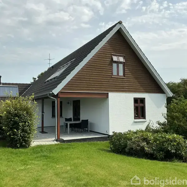 Et enkelt, hvidt hus med en træbeklædt gavl står i en velplejet have. Der er havemøbler på en overdækket veranda, omgivet af grønne buske og græs.