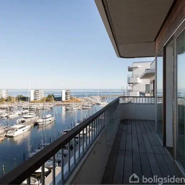 En altan med glasrækværk strækker sig ud fra en lejlighed, med udsigt til en marina fyldt med sejlbåde, omgivet af moderne lejligheder og havet i horisonten.