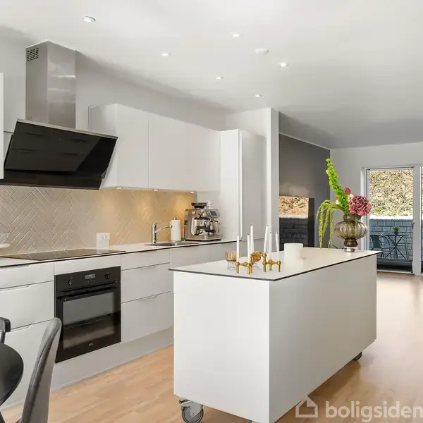 Et moderne køkken med hvide skabe og en sort kogeplade. Et centralt køkkenbord står foran, dekoreret med lysestager. Store glasdøre fører til en balkon med udsigt over grønt område.