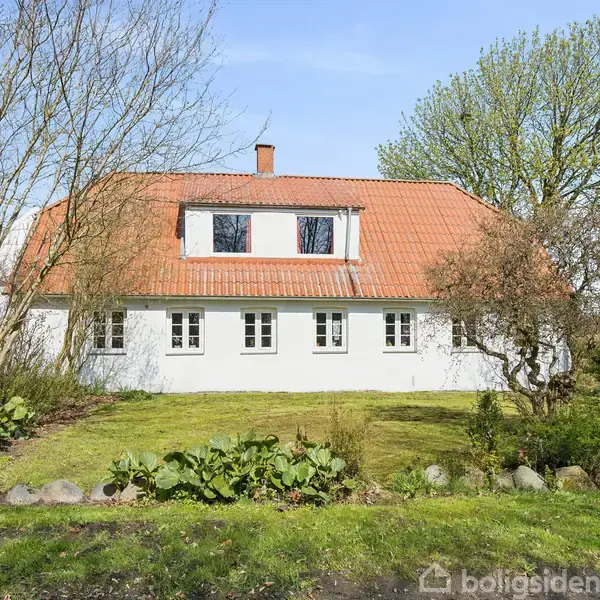 Et hvidt hus med rødt tegltag står på en grøn græsplæne omgivet af forskellige træer og buske. Huset har flere vinduer og en centralt placeret skorsten.