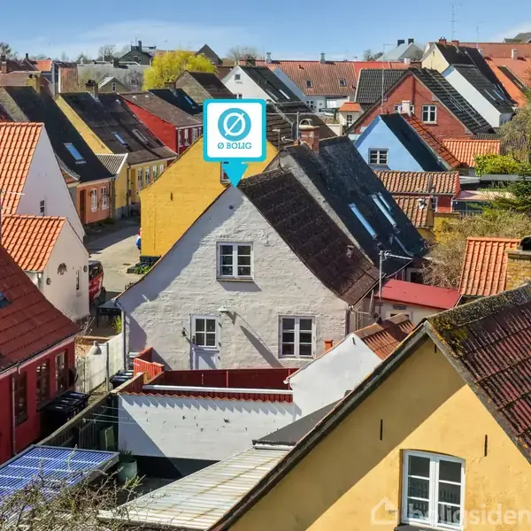 Et rækkehus med røde tegltage ligger i en lille by med smalle gader og farverige bygninger. Et skilt på taget viser teksten "0 BOLIGD".
