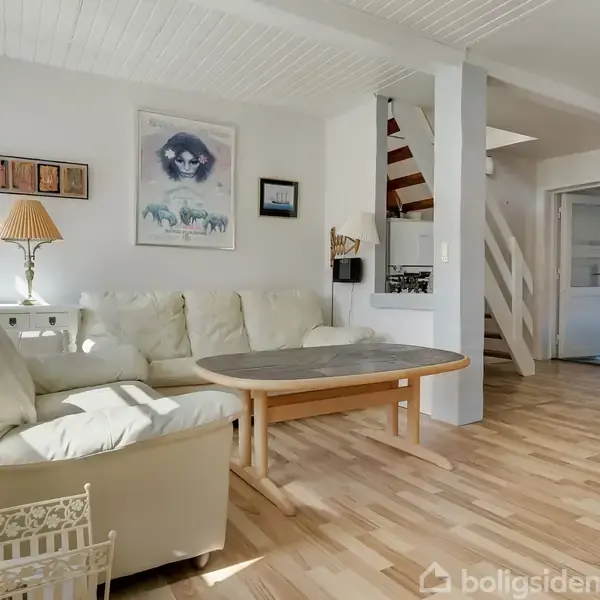 En beige sofa står i en lys stue med trægulv, hvor et rundt træbord er placeret foran. Der er malerier og en lampe i baggrunden ved væggen.