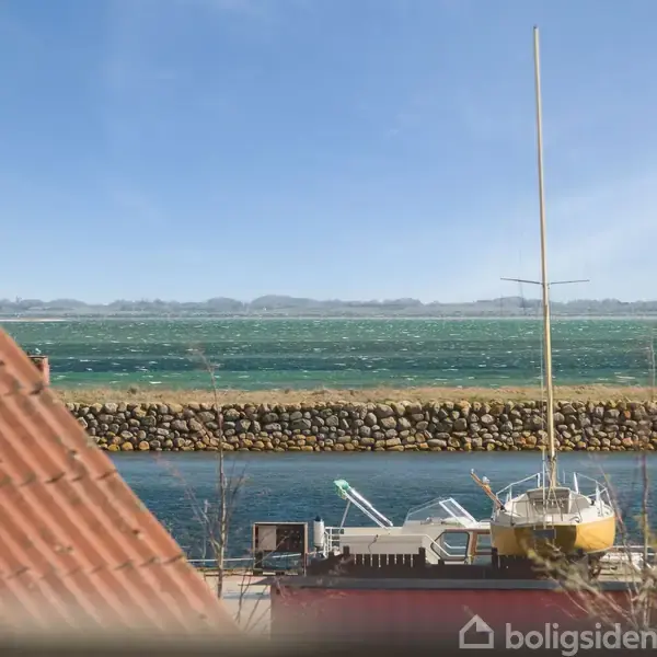 En sejlbåd ligger stille ved en kaj. Bag den ses blåt vand og en stenmur i baggrunden. Røde og blå bygninger er synlige i forgrunden.
