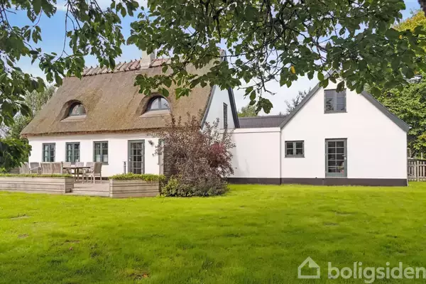 Et hvidt stråtækt hus står stille i en grøn have med en terrasse og træmøbler. Træer omkranser området under en klar blå himmel.