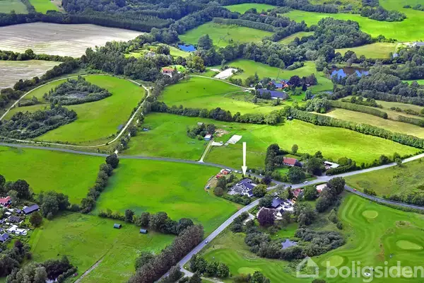 Luftfoto af frodige, grønne marker med spredte gårde og huse. Flere veje snor sig gennem landskabet, som er omkranset af skove og skoveområder.