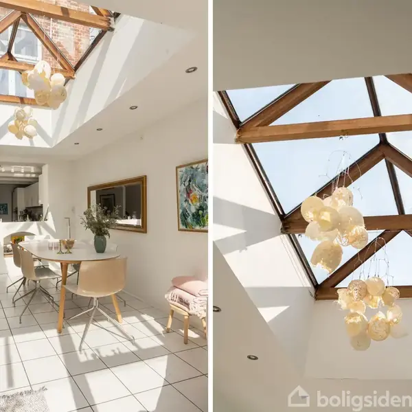 Et lyst køkken-alrum med hvide fliser og en lang spisebord i fokus, oplyst af naturligt lys fra et stort ovenlysvindue med træbjælker. En lysekrone hænger fra loftet.