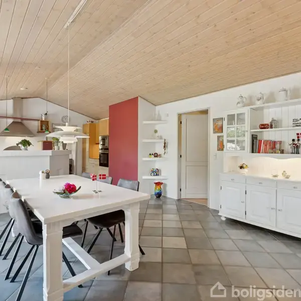 Et køkken med trækabinetter og grå kogeplader under en emhætte. Et vindue lukker naturligt lys ind, mens en dør fører til en gang med flisegulv. En kande står på en hylde.