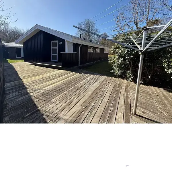 Et sort træhus står på en stor terrasse af træplanker. En tørrestativ er placeret i terrassen. Træer og buske omkranser området, og himlen er klar og blå.