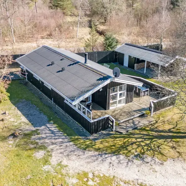 Sort hus med terrasse set fra oven, beliggende i et skovområde. Terrassen er omgivet af et hegn, og en garage står lidt væk fra huset. Solbeskinnet landskab.