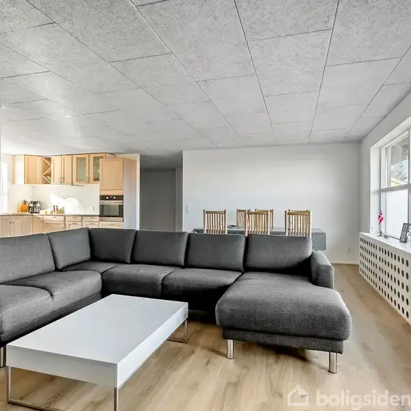 Grå sofa står i hjørne, omkring et hvidt sofabord. Baghus ses et spisebord med fire stole. Store vinduer til højre slipper lys ind i det moderne rum.