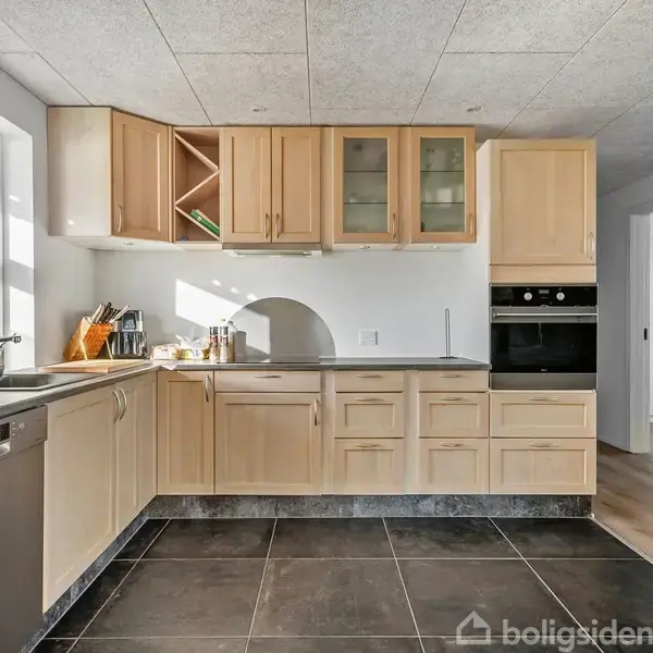 Køkken med lyse træskabe og sort bordplade. Opvaskemaskine og vask er indbygget. Store fliser på gulvet. Inventar omfatter køkkenredskaber ved vasken. Lyse vægge og lofter, dør til højre.