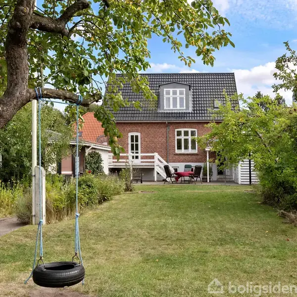 Et rødt murstenshus med sort tegltag står i baggrunden. En græsplæne med et dækgyngestativ i et træ dominerer forgrunden. Terrasse med havemøbler ses foran huset.