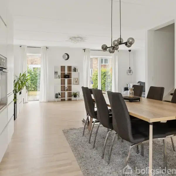 Spisebord med otte stole står på et tæppe. Bagved er et reolsystem ved vinduer med gardiner, planter og dekorationer. Moderne lamper hænger fra loftet i et lyst rum.