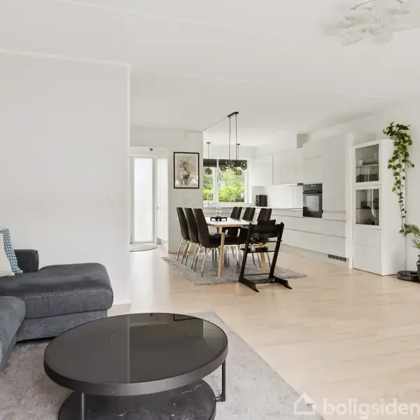 En moderne stue med en grå sofa og et rundt sort sofabord, står ved siden af en spisestue med sort bord og stole. Et hvidt køkken ses i baggrunden.