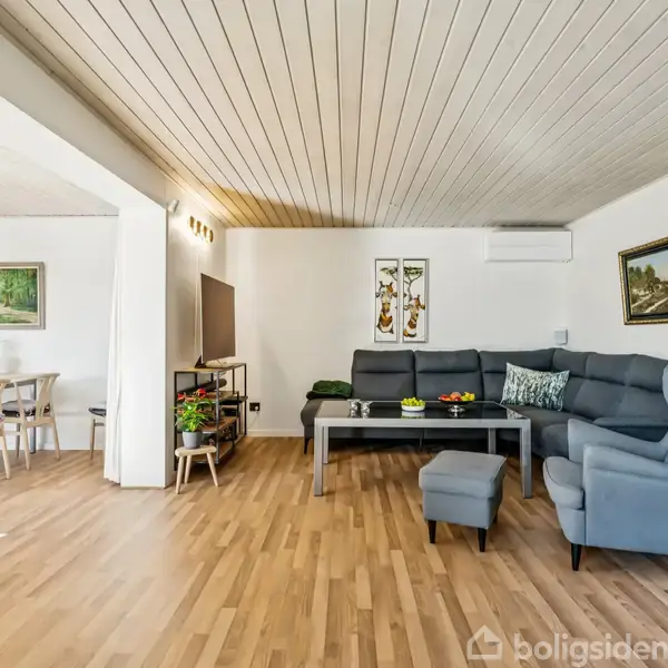 Stue med gråt hjørnesofaarrangement, der står foran et mørkebord med dekorationer; spisestue med træmøbler i baggrunden, vægmalerier og en lysende lampe skaber en hyggelig atmosfære.