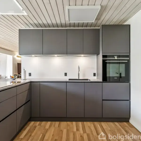 Moderne køkken med mørkegrå skabe og indbygget ovn, centreret omkring en hvid bordplade. Rummet har lyst trægulv og et hvidt loft med nedsænkede paneler.