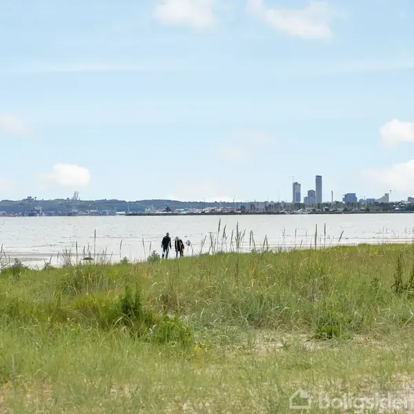 Byggeplads med fundament i forgrunden, omgivet af græs og buske. Bagved ses en strand med havudsigt og en klar blå himmel. En enkeltstående træ til venstre.